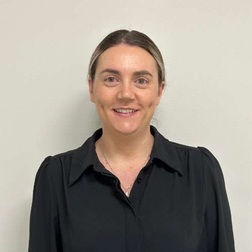 A woman with brown hair stands against a plain backdrop, wearing a black blouse and a necklace, smiling at the camera. A woman with brown hair stands against a plain backdrop, wearing a black blouse and a necklace, smiling at the camera.