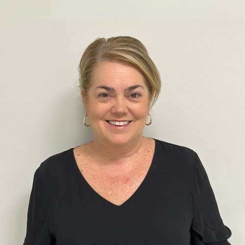 A person with short hair wearing a black top smiles while standing against a plain white background. A person with short hair wearing a black top smiles while standing against a plain white background.