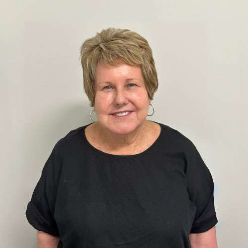 5 A woman with brown hair stands against a plain backdrop, wearing a black blouse and a necklace, smiling at the camera.