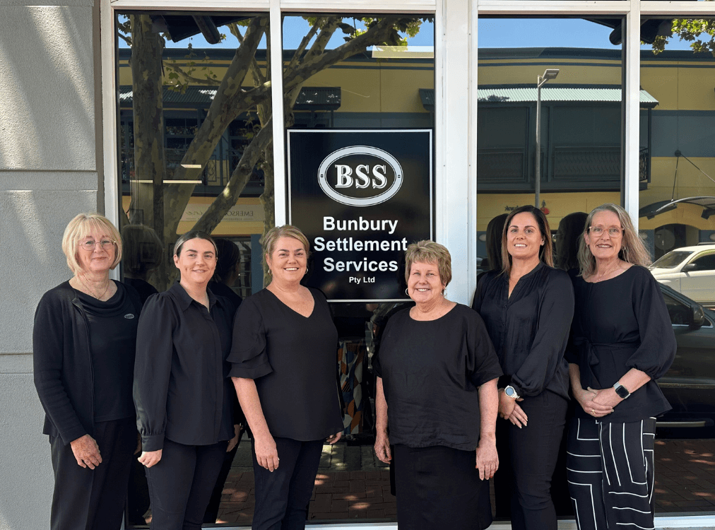 Six women standing in front of a business entrance with a sign that reads "Bunbury Settlement Services Pty Ltd." They are all wearing black clothing. Six women standing in front of a business entrance with a sign that reads "Bunbury Settlement Services Pty Ltd." They are all wearing black clothing.