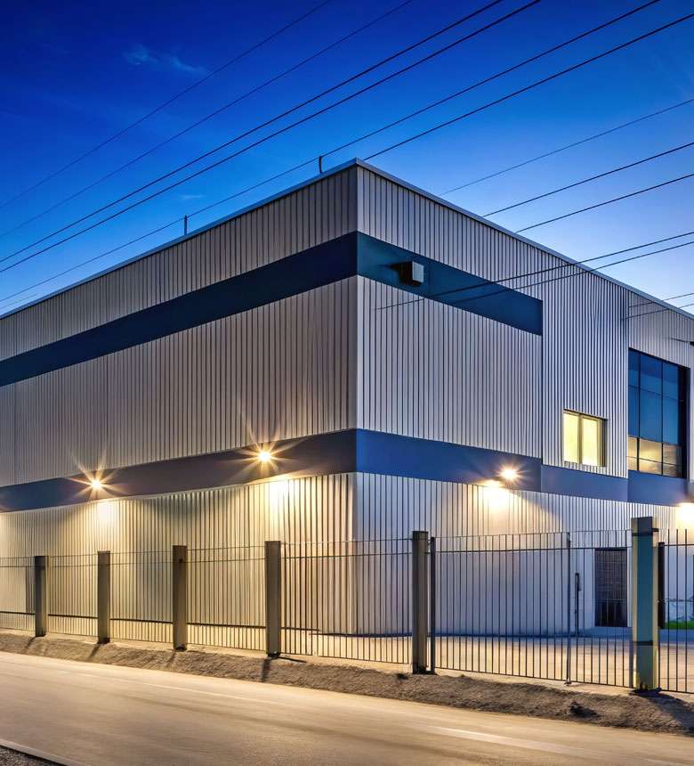 Industrial building at dusk, illuminated by exterior lights, with a metal fence in the foreground and overhead power lines. Industrial building at dusk, illuminated by exterior lights, with a metal fence in the foreground and overhead power lines.