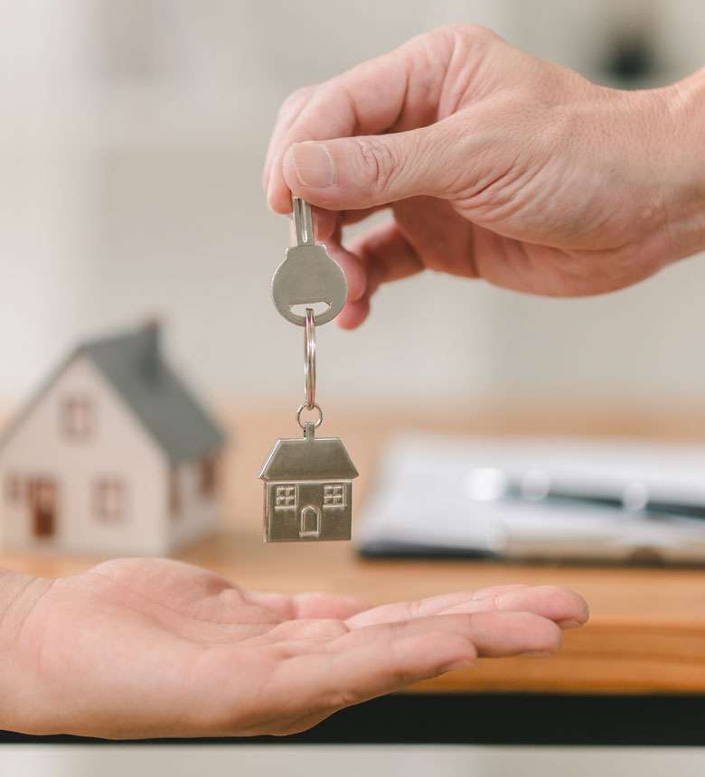 A hand giving a house-shaped keychain to another open hand. A small model house is in the background on a table.