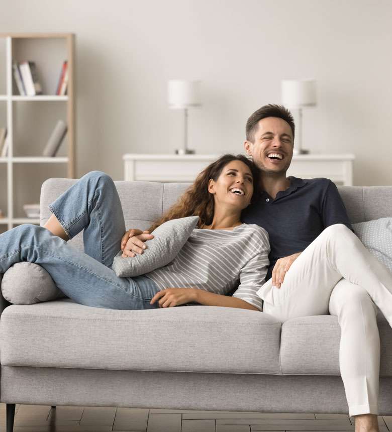 A couple sits on a gray sofa in a living room, laughing and holding a pillow. Shelves and lamps are visible in the background. A couple sits on a gray sofa in a living room, laughing and holding a pillow. Shelves and lamps are visible in the background.