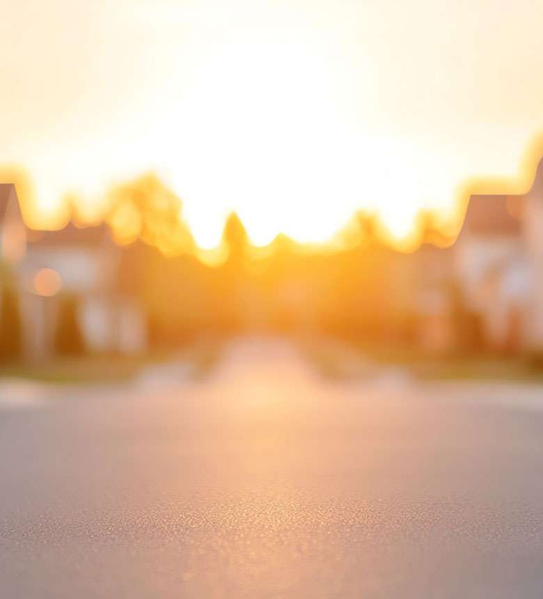 Blurred view of a sunlit neighborhood street at sunrise or sunset, with houses and trees in the background.