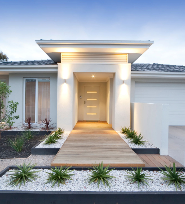 Modern house exterior with a wooden pathway leading to a front door, flanked by plants and white stone landscaping under a clear sky. Modern house exterior with a wooden pathway leading to a front door, flanked by plants and white stone landscaping under a clear sky.